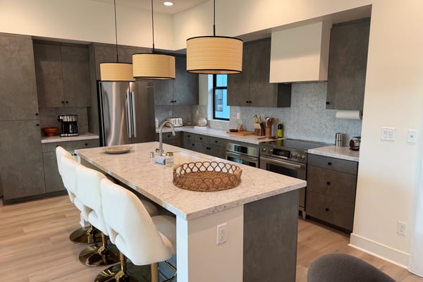 Modern kitchen with dark gray cabinetry, marble countertops, stainless steel appliances, large island with white seating, and pendant lights overhead.