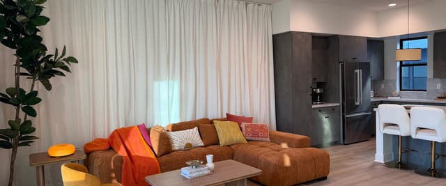 Open-plan living room with brown sectional sofa, yellow accent chair, wooden coffee table, and kitchen with dark cabinetry visible in background