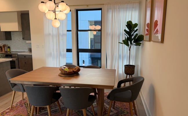 Modern dining room with wood table, gray chairs, pendant lighting, and potted plant near floor-to-ceiling windows
