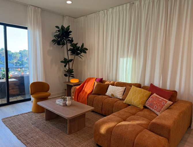Modern living room with brown sectional sofa, colorful pillows, wooden coffee table, orange accent chair, potted plant, and floor-to-ceiling cream curtains